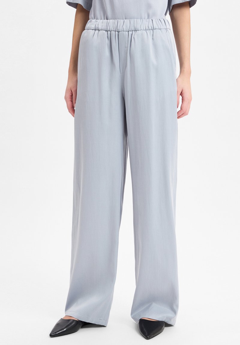 Vrouw in lichtgrijze wijde broek met hoge taille en elastische tailleband, met zwarte puntige ballerina's, op een witte achtergrond.