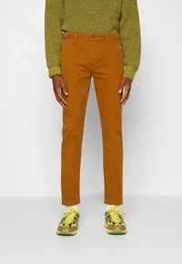 Pantalon chino en coton marron coupe slim, associé à des chaussures de sport jaunes vives et un pull vert texturé.