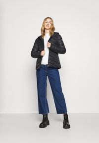 Čierna prešívaná puffová bunda s kapucňou, predným zipsom a pruhovaným vzorom na rukávoch. V kombinácii s voľnými modrými džínsami a čiernymi členkovými čižmami.