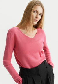 Różowy sweter w serek wykonany z miękkiego materiału z długimi rękawami, zestawiony z czarnymi, plisowanymi spodniami o wysokim stanie. Gładka tekstura i dopasowany krój.