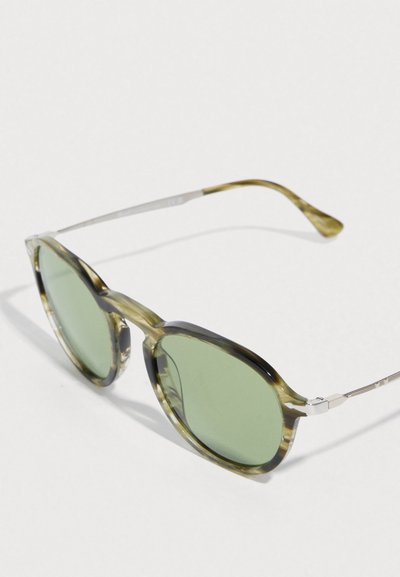 Πράσινα γυαλιά ηλίου με οκταγωνικό σχήμα, διαθέτοντας διαφανές πράσινο και μαύρο σκελετό και ασημένιους μεταλλικούς βραχίονες.