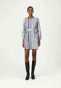 Naine, kellel on seljas tükkide pikkune valge kleit, mis on vertikaalsete siniste triipudega ja vööga, ning mustad põlve kõrgused saapad, seisab valge tausta ees.