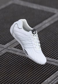 Hvid sportsko med et slankt design, læderoverdel, seks perforerede striber, snørebåndslukning og et sort Adidas-logo på tungen.