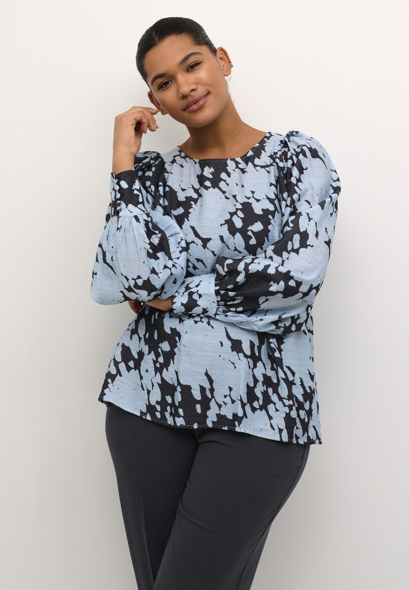 Blaue gemusterte Bluse mit schwarzem abstraktem Design, Rundhalsausschnitt, langen Puffärmeln und lässiger Passform, kombiniert mit dunklen Hosen.