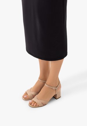 Piedi che indossano sandali beige con tacco a blocco e cinturini alla caviglia, abbinati a una gonna midi nera, mostrati su sfondo bianco.
