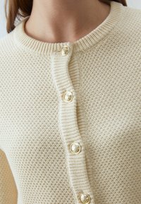 Kremowy sweter z dzianiny o teksturowej powierzchni, z prążkowanym kołnierzem i dużymi złotymi guzikami z przodu.