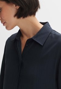 Navyblaues Hemd aus strukturiertem Stoff, mit einem spitzen Kragen und Knopfdetails, seitlich gezeigt mit sichtbarem kurzen Haar.