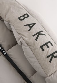 Beige gesteppte Jacke mit großen schwarzen "BAKER" Buchstaben auf dem Kragen, die aus glattem Stoff und gepolsterter Textur besteht. Schwarzer Reißverschluss als Akzent.