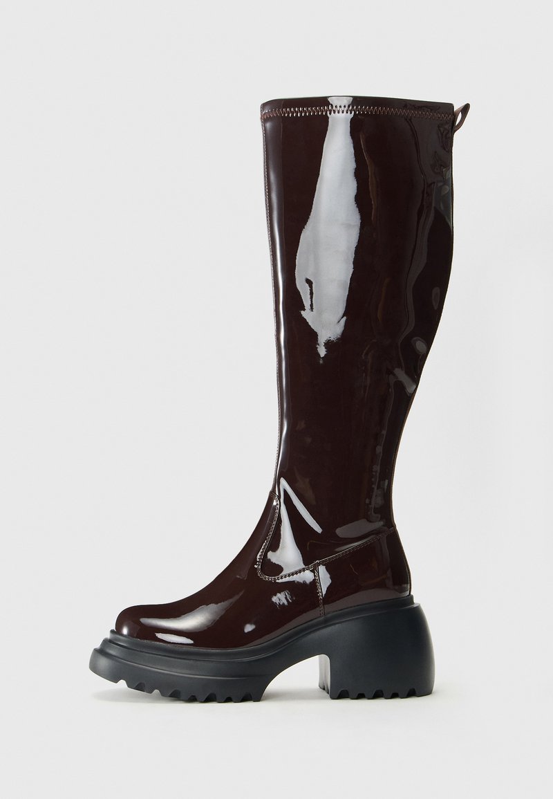 Térdig érő, burgundi színű, fényes gumicsizma vastag fekete platform talppal és textúrázott talppal, látható varrószegésekkel és húzófüllel.