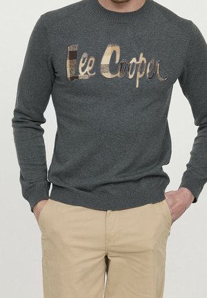 Mężczyzna w ciemnoszarym dzianinowym swetrze z beżowym kratkowanym logo "Lee Cooper", beżowych spodniach, ręce w kieszeniach, stoi na jednolitym tle.