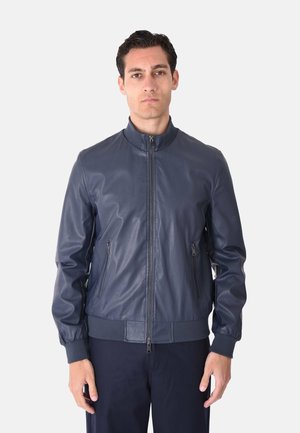 Uomo in piedi che indossa una giacca di pelle blu navy con cerniere laterali e polsini e orlo a coste.