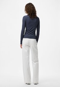 Mulher com cabelo encaracolado, vestindo uma blusa justa de mangas longas escuras e calças brancas largas, de pé, de costas, num fundo branco liso.