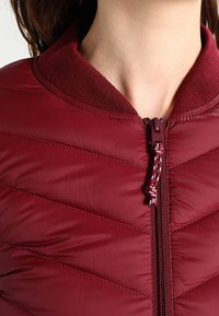 Burgundy kauluspaita, jossa on teksturoidut, uritetut kaulukset ja erivärinen vetoketjun vetokappale. Kirjailuja on diagonaalisesti, mikä tarjoaa lisäeristyksen.