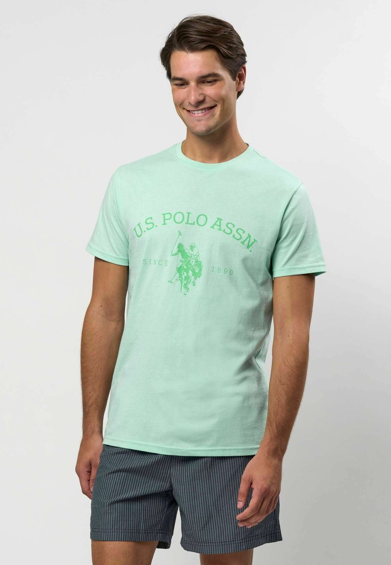 Jasnozielona bawełniana koszulka z okrągłym dekoltem i przednim graficznym nadrukiem w ciemniejszym odcieniu zieleni, zestawiona z ciemnymi spodniami w paski.