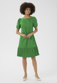 Zelena bombažna obleka s kratkimi nabranimi rokavi in plisejem na robu. Ima kvadratni izrez in sega nekoliko nad kolena. Postavljena na nevtralno ozadje.