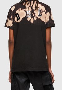 Černé bavlněné tričko s designem posetým bělicími skvrnami, s tlustým bílým nápisem "DSL" na zadní straně. Krátké rukávy a volný střih.