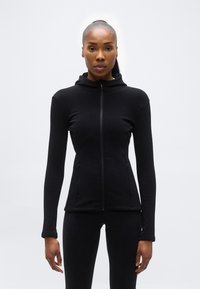 Γυναίκα που στέκεται και κοιτάζει μπροστά, φορώντας μια εφαρμοστή μαύρη ζακέτα με φερμουάρ και κουκούλα και αντίστοιχο μαύρο κολάν, με φόντο ένα απλό φωτεινό υπόβαθρο.