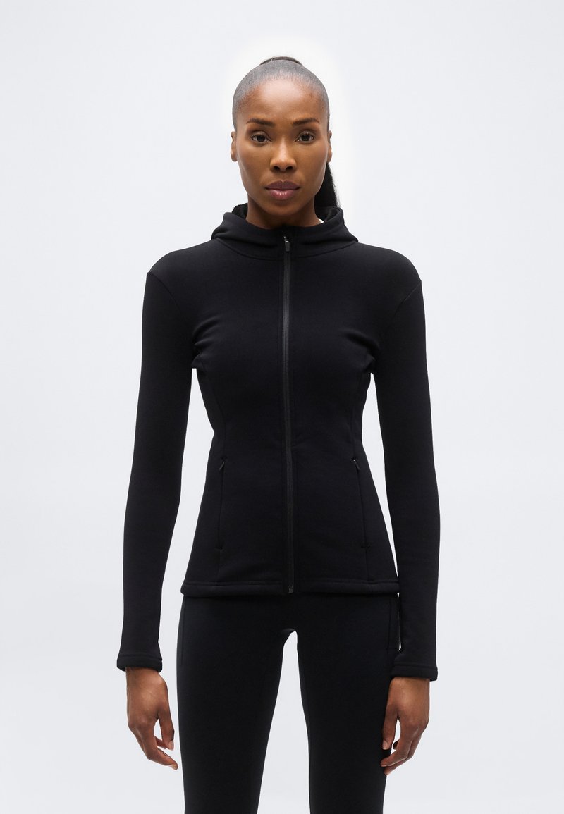 Γυναίκα που στέκεται και κοιτάζει μπροστά, φορώντας μια εφαρμοστή μαύρη ζακέτα με φερμουάρ και κουκούλα και αντίστοιχο μαύρο κολάν, με φόντο ένα απλό φωτεινό υπόβαθρο.