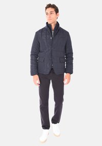 Giacca imbottita blu navy con due tasche frontali, chiusura a bottoni e colletto alto; indossata sopra un maglione grigio e abbinata a pantaloni scuri e sneakers bianche.