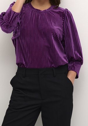 Femme portant un chemisier en velours côtelé violet à manches longues, rentré dans un pantalon noir taille haute, une main dans la poche.