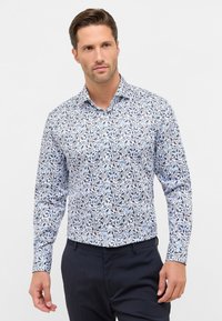 Mann trägt ein eng anliegendes, langärmliges weißes Hemd mit blauem und schwarzem Blumenmuster, kombiniert mit dunkelblauen Hosen, steht vor einem weißen Hintergrund.