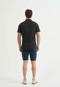 Chemise noire à manches courtes avec un ourlet arrondi, associée à un short bleu marine et des chaussures claires, vue de dos.