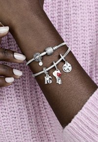 Srebrni čarovni zapestnice z raznolikimi oblikami, ki vsebujejo teksturirane perle in čare, vključno s srčki in črkami, na zapestju, ki je oblečeno v roza pulover.