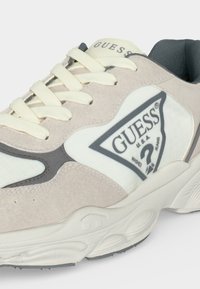 Baskets Guess beige clair et blanches avec des lacets crème, une semelle épaisse, et le logo Guess sur le panneau latéral et la languette.