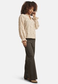 Beige floral-embroidered blouse with long sleeves and ruffled cuffs, paired with brown checked flared trousers and beige heeled boots.