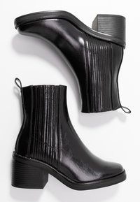 Par de botas de tobillo de cuero negro con tacones cuadrados y paneles laterales elásticos, mostradas sobre un fondo blanco.