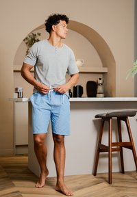 Camiseta henley de mangas cortas gris y pantalones cortos a rayas azul claro, de pie descalzo en una cocina con detalles de madera y electrodomésticos modernos.