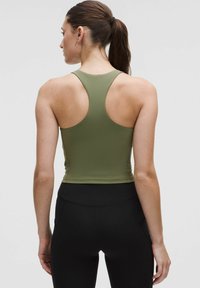 Donna con coda di cavallo castana che indossa un top corto a canottiera verde oliva e leggings neri a vita alta, in piedi davanti a uno sfondo neutro.