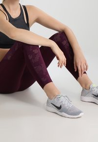 Reggiseno sportivo nero e leggings bordeaux con motivi testurizzati, abbinati a scarpe da corsa grigie con un baffo nero e accenti neon.