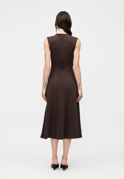 Γυναίκα με μακριά σκούρα μαλλιά, που φοράει ένα αμάνικο καφέ μίντι φόρεμα και μαύρα παπούτσια με τακούνι, στέκεται με την πλάτη σε μονόχρωμο φόντο.