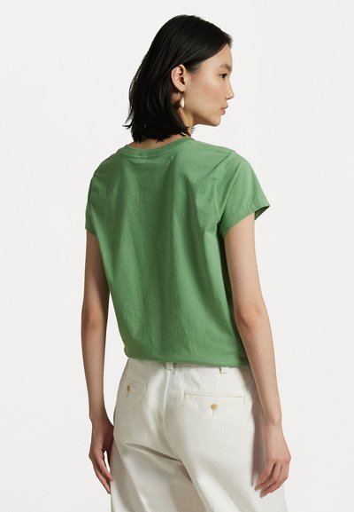 Green cotton t-shirt with short sleeves, round neckline, and a relaxed fit. Worn with light-colored trousers, showcasing a smooth texture.