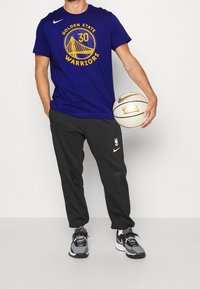 Modré tričko s logem "Golden State Warriors" v žluté barvě a černé tepláky; drží basketbal; značka Nike na oblečení a botách.