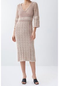 Robe en maille beige clair, avec col en V, manches trois-quarts, motif ajouré, et longueur jusqu'aux genoux. Associée à des mules noires.