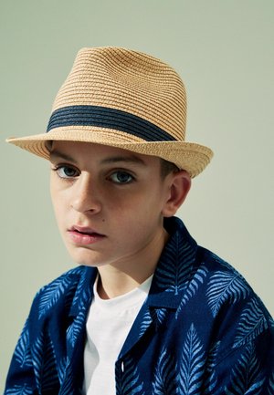 Geflochtener Strohhut in hellem Beige mit einem dunkelblauen Band; kombiniert mit einem blauen Hemd mit einem Blattmuster über einem weißen T-Shirt.