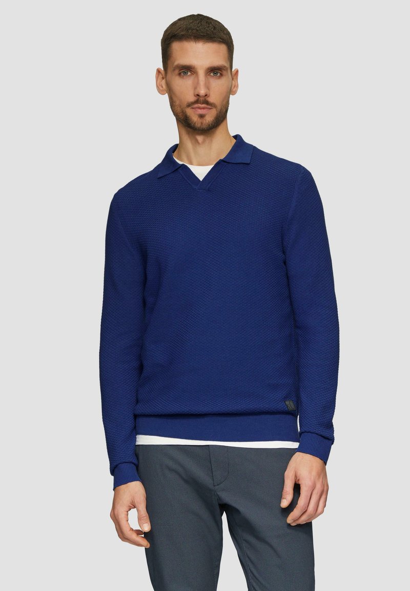 Marineblauer Pullover mit strukturiertem Strickmuster, V-Ausschnitt und gerippten Bündchen. Über einem weißen Hemd getragen.