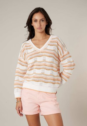 Femme portant un pull en maille à rayures blanches et pastel orange avec un col en V et un short rose pâle retroussé, debout avec une main sur la hanche.