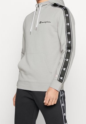 Homme portant un sweat à capuche Champion gris clair avec un ruban noir à logo sur les manches et un pantalon noir assorti avec un ruban similaire sur le côté.