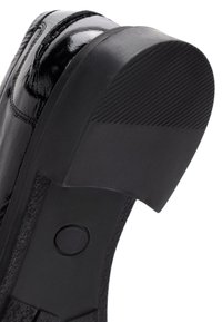 Zapato de charol negro con punta redonda y suela de goma negra texturizada. Presenta un acento circular cerca de la base.