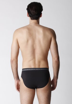 Uomo in piedi di spalle, indossa slip neri con fascia in vita con marchio, su sfondo chiaro uniforme.