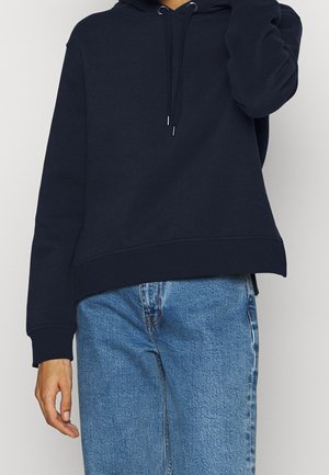 Person trägt einen schlichten schwarzen Hoodie mit Kordelzug und hellblaue, hoch taillierte Jeans, Hand ruht nahe der Hüfte.