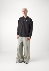 Zwarte pullover met lange mouwen en een half-rits kraag, voorzien van een logo; gecombineerd met lichtgrijze baggy cargo pants met zijzakken en een relaxte pasvorm.