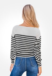 Mujer con cabello largo y rubio viste un suéter de rayas blanco y negro y jeans azules, de pie de espaldas contra un fondo blanco.