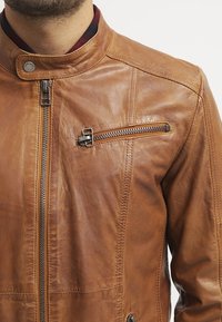 Brun læderjakke med opretstående krave, lynlåslukning foran og sidelommer. Har en diagonal lynlåslomme på brystet. Glat tekstur.