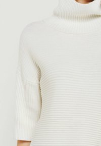 Weißer gerippter Pullover mit hohem Kragen, kurzen Ärmeln und einer weichen Textur, der ein dezentes vertikales Muster aufweist.