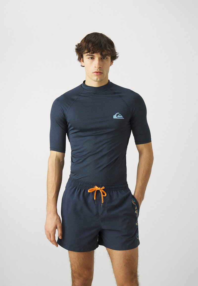 Navy lyhythihainen rash guard sinisellä logolla, yhdistettynä tummansinisiin uimapöksyihin, joissa on oranssi naru ja kuvioidut sivuakkentit.