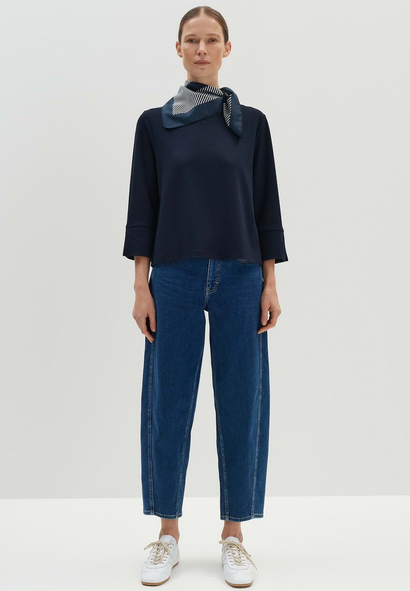 Haut à manches longues bleu marine avec des manches évasées, associé à un jean en denim bleu taille haute et des baskets blanches. Écharpe à rayures bleu marine et blanches.